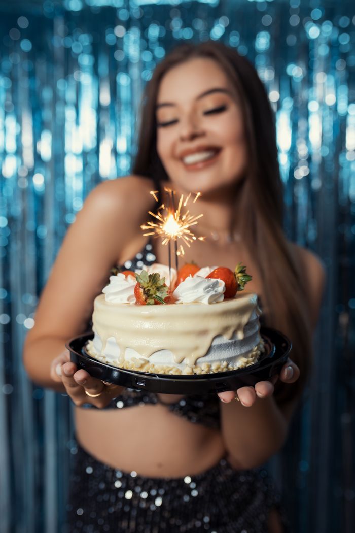 A happy young woman holds a cake with a sparkler against a festive backdrop, celebrating a birthday in Brazil.